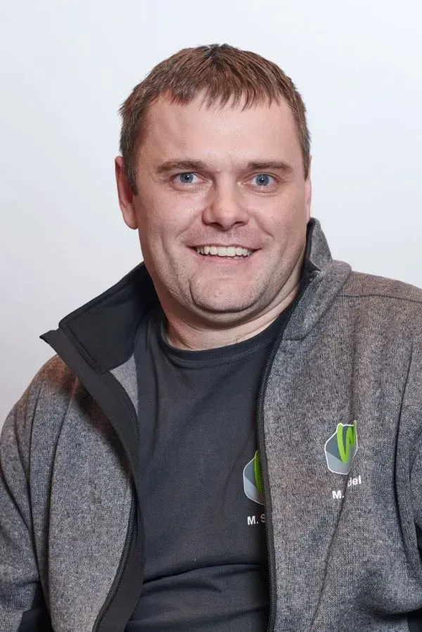 Ein Mann in einer grauen Jacke und einem schwarzen Hemd lächelt in die Kamera.