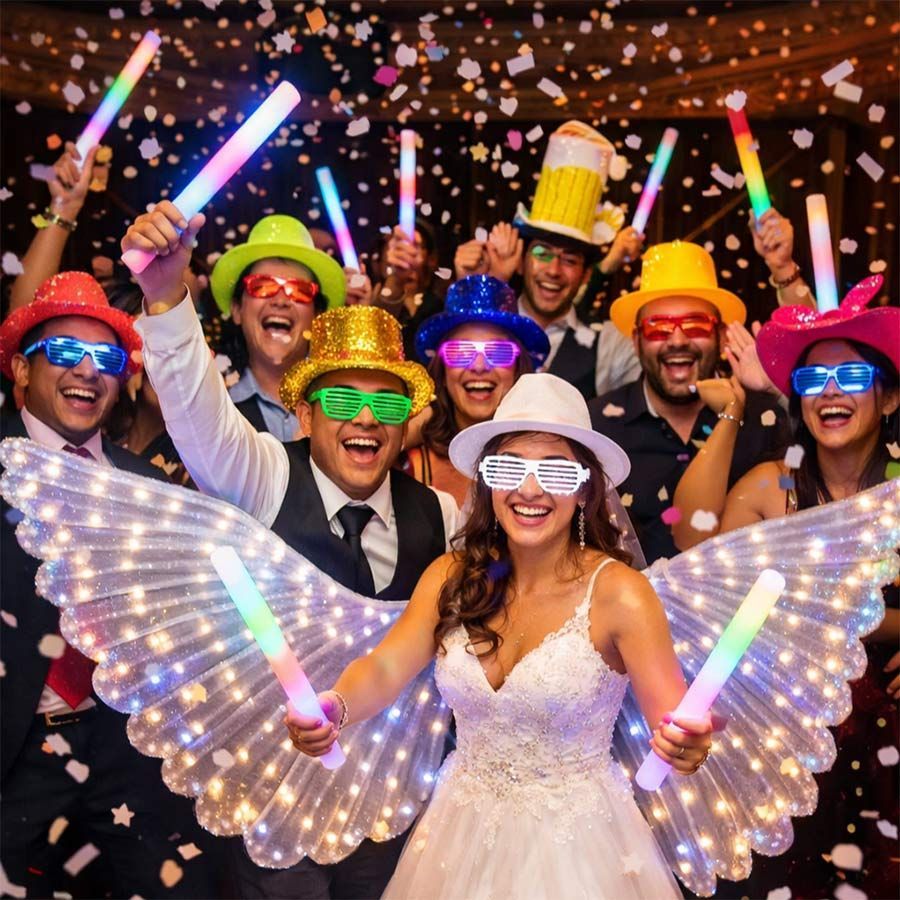 Grupo de personas con sombreros de fiesta, gafas y barras luminosas celebrando con confeti. Una mujer con vestido blanco tiene alas luminosas.