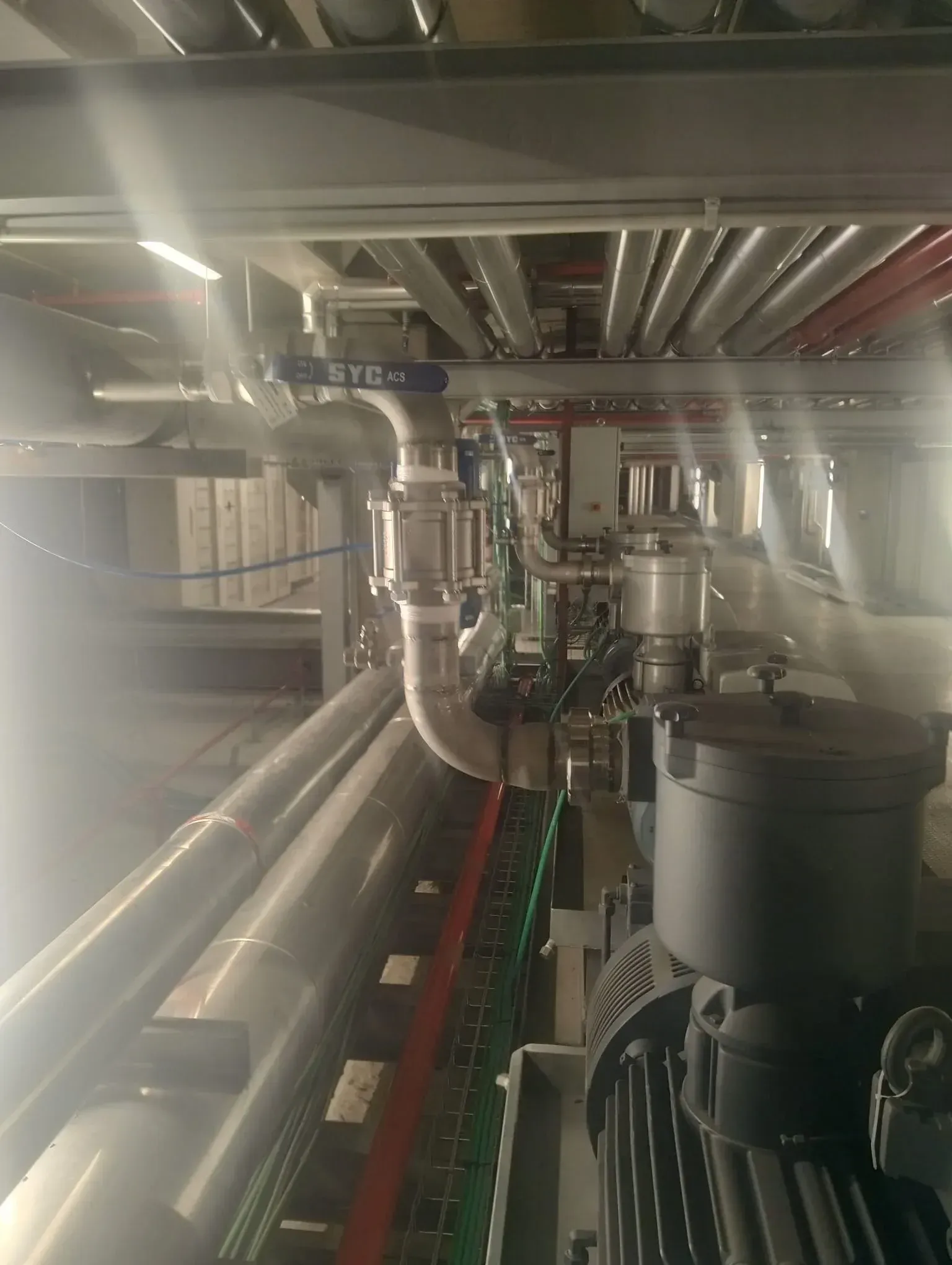 Tuberías y conductos industriales dentro de una sala de máquinas metálica, con tuberías rojas y grises en el techo.