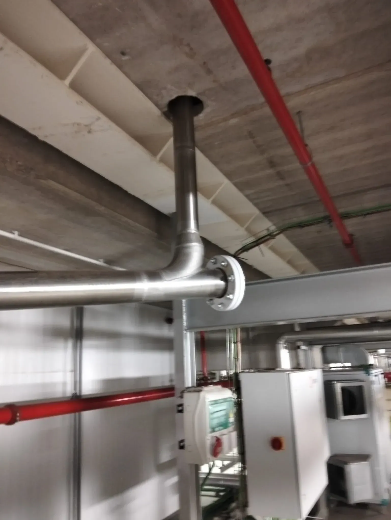 Techo industrial con tuberías plateadas y conductos rojos sobre equipos en un cuarto de servicio.