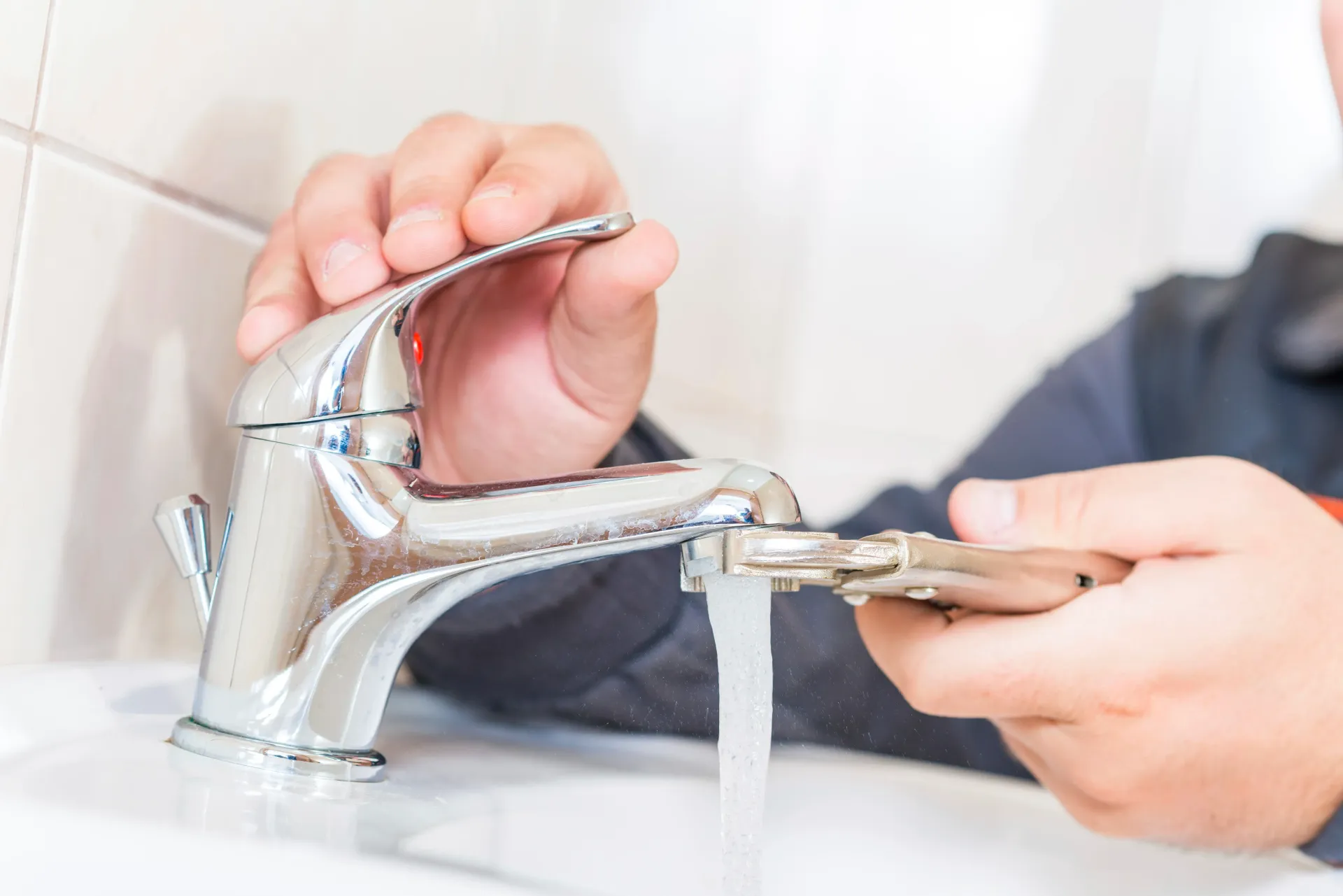 Manos usando una llave inglesa para apretar un grifo cromado con agua corriendo en un lavabo de baño.