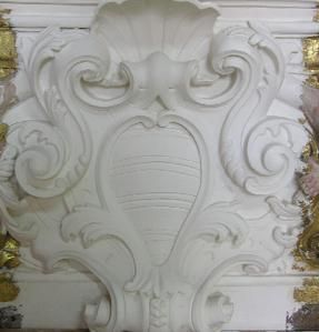 Gesso branco ornamentado de uma urna e folhas em espiral, possivelmente um detalhe arquitetônico, com detalhes dourados.