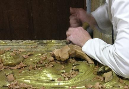 Uma pessoa vestida de branco esculpindo madeira com um cinzel, superfície dourada com aparas de madeira.