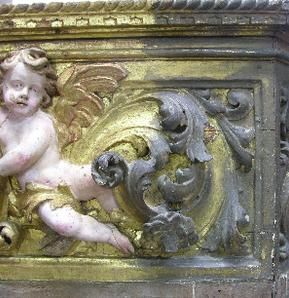 Um querubim e um arabesco ornamentado em dourado e cinza sobre uma superfície de madeira desgastada.