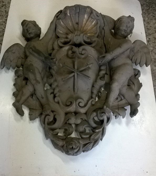 Relevo esculpido em madeira com dois querubins ladeando um escudo com um símbolo de cruz e uma guirlanda de flores.