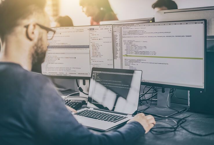 Eine Person arbeitet an einem Schreibtisch mit mehreren Computermonitoren, auf denen Code angezeigt wird, in einem hellen Büro.