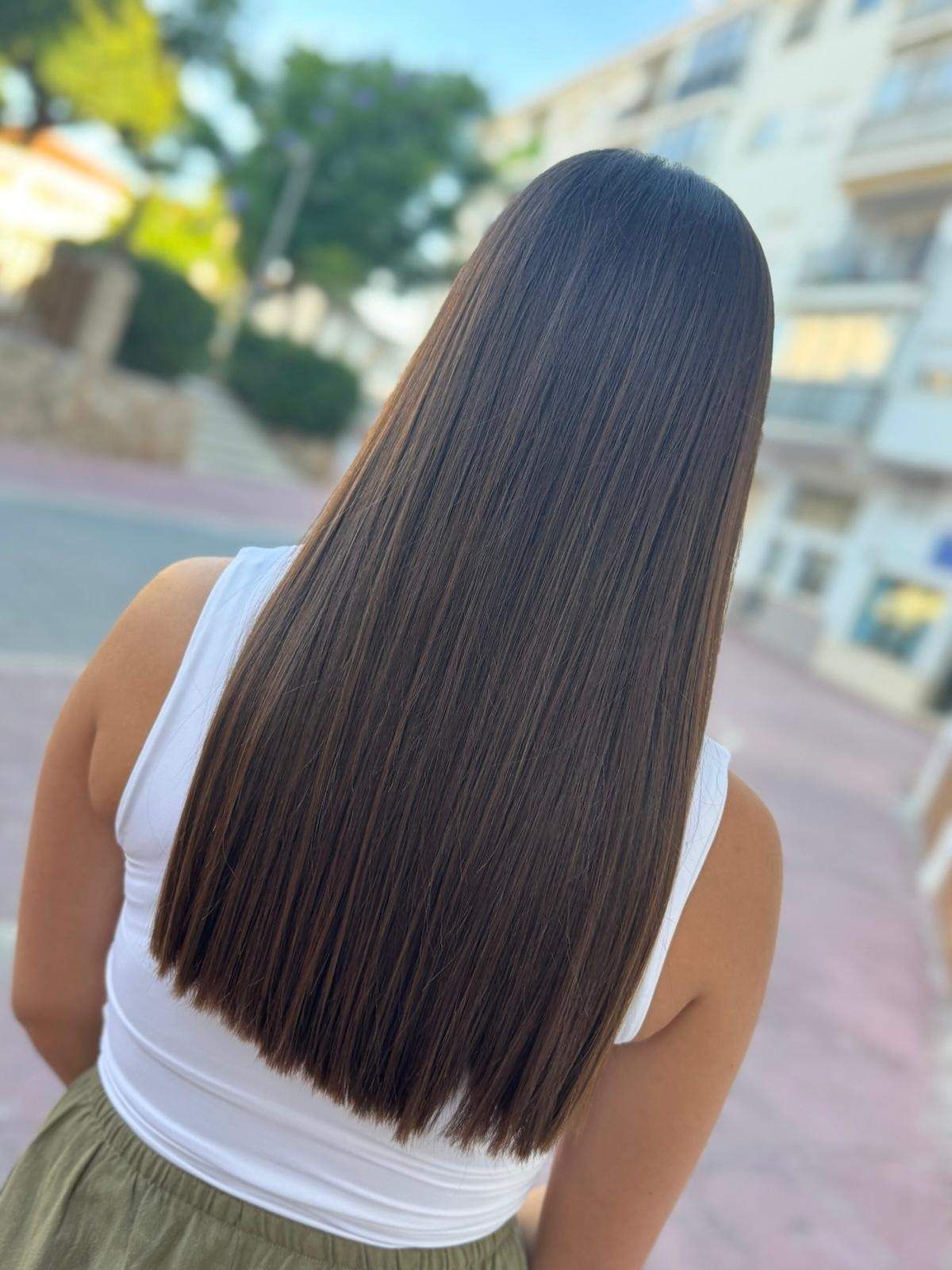 Vista posterior de una persona con micro trenzas largas, rectas y de color marrón oscuro, vestida con una blusa blanca y una falda verde, al aire libre.
