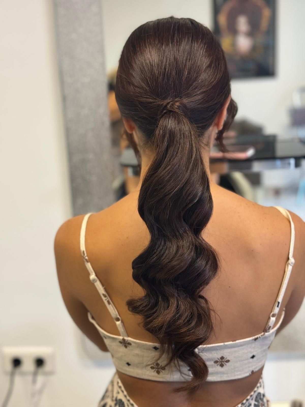 Mujer con cabello castaño oscuro peinado en una cola ondulada, vistiendo un top con estampado floral blanco.