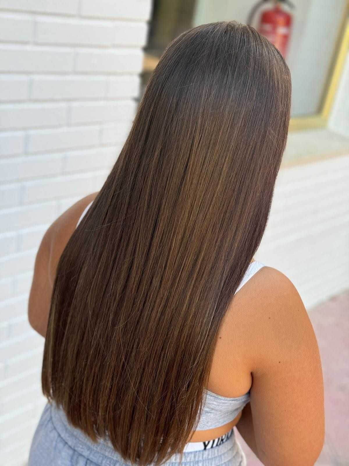 Mujer de espaldas con microtrenzas largas y rectas de color marrón. Lleva una blusa blanca y está de pie junto a una pared de ladrillos blancos.