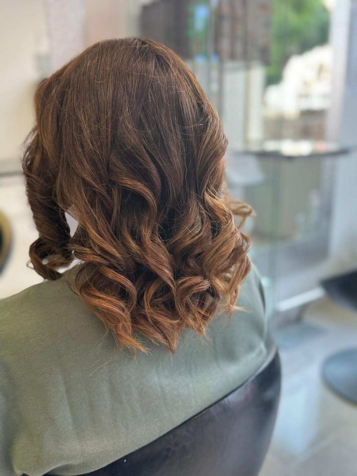 Mujer con cabello castaño ondulado peinado con rizos, sentada en una silla de salón.