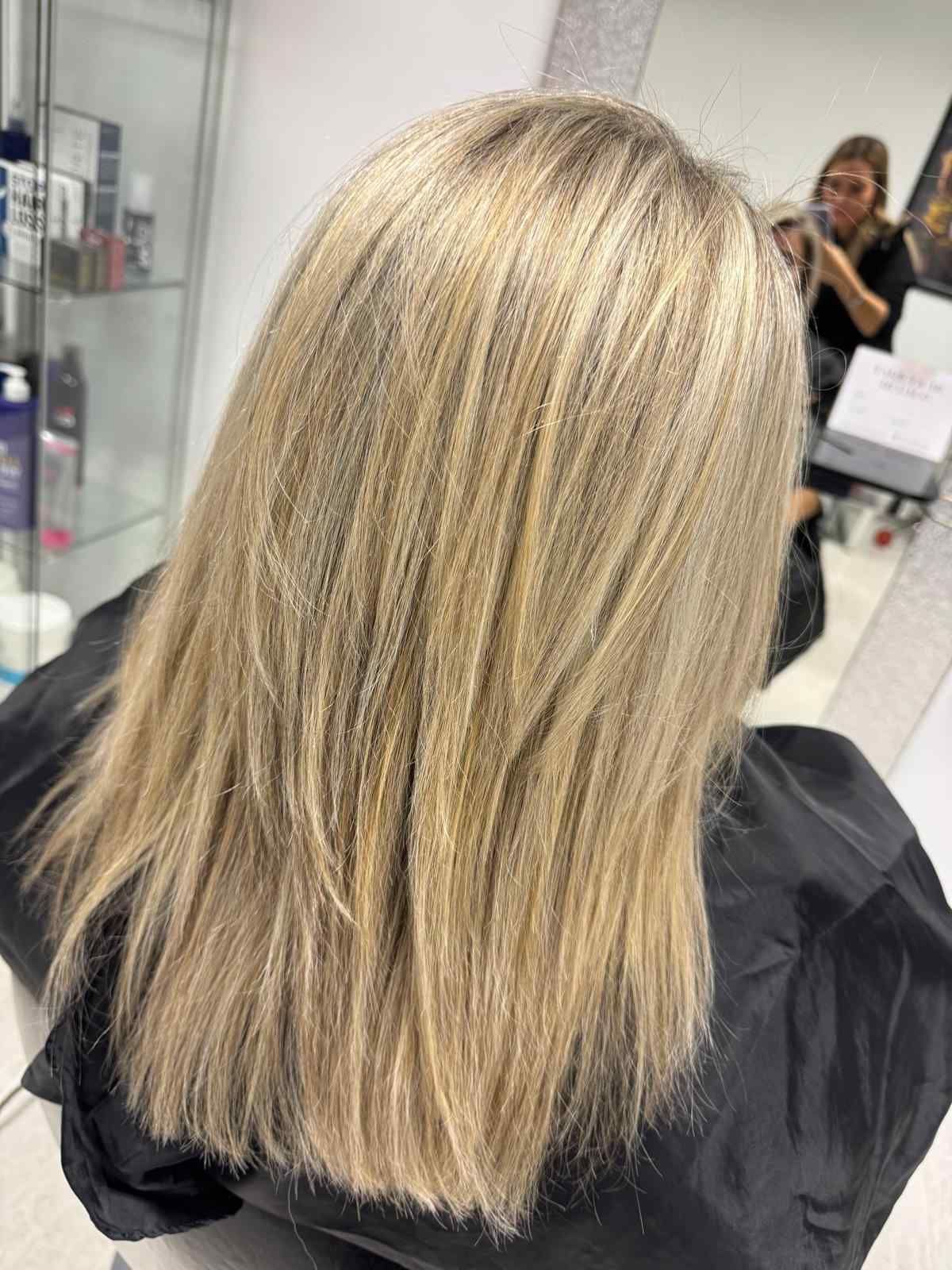 Cabello rubio peinado con capas, estilo salón. Se ve a una estilista en el espejo.