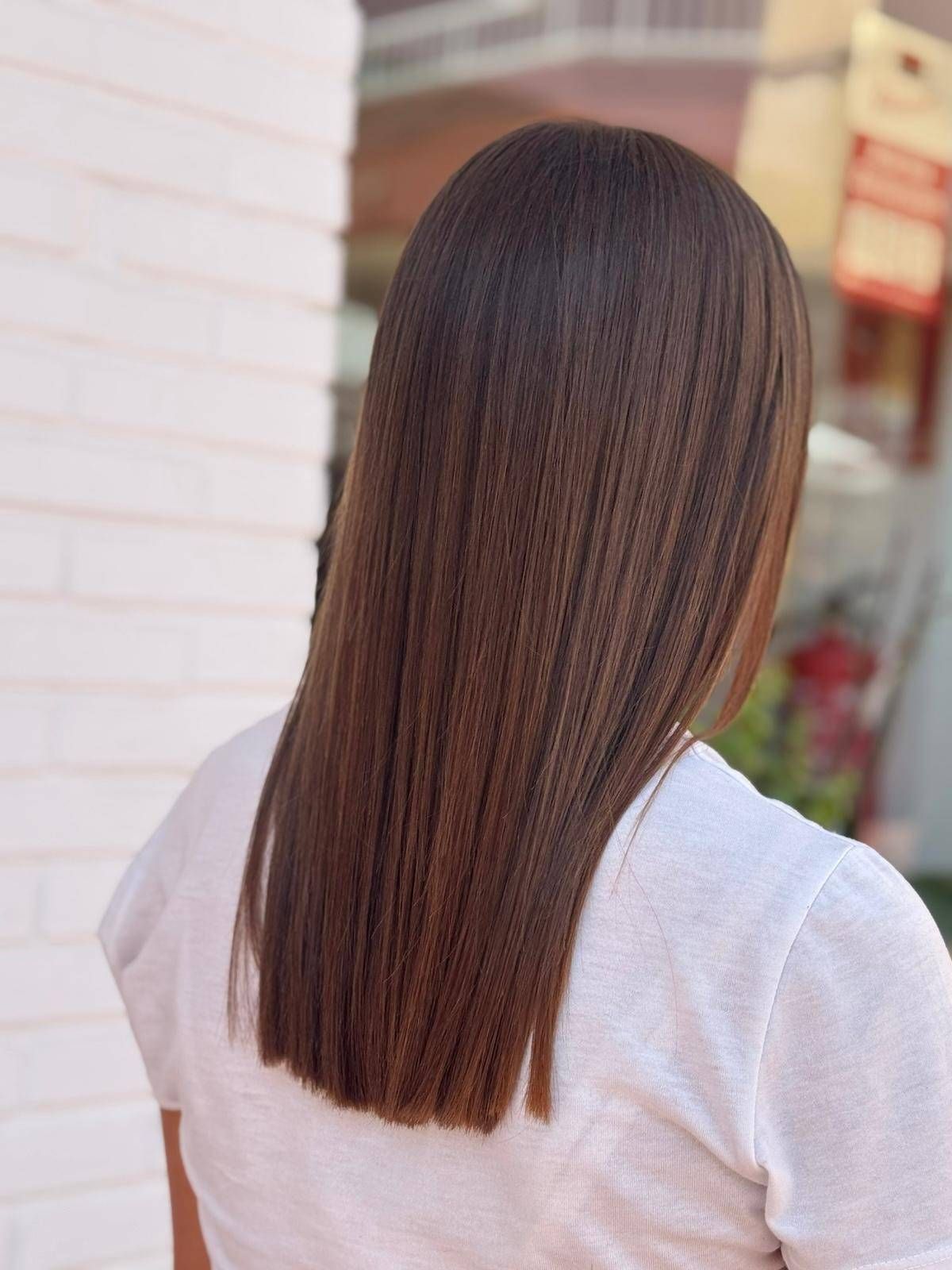 Cabello castaño largo, liso y rizado de mujer, hasta los hombros, con una parte superior texturizada de color blanco, contra una pared de ladrillos blancos.