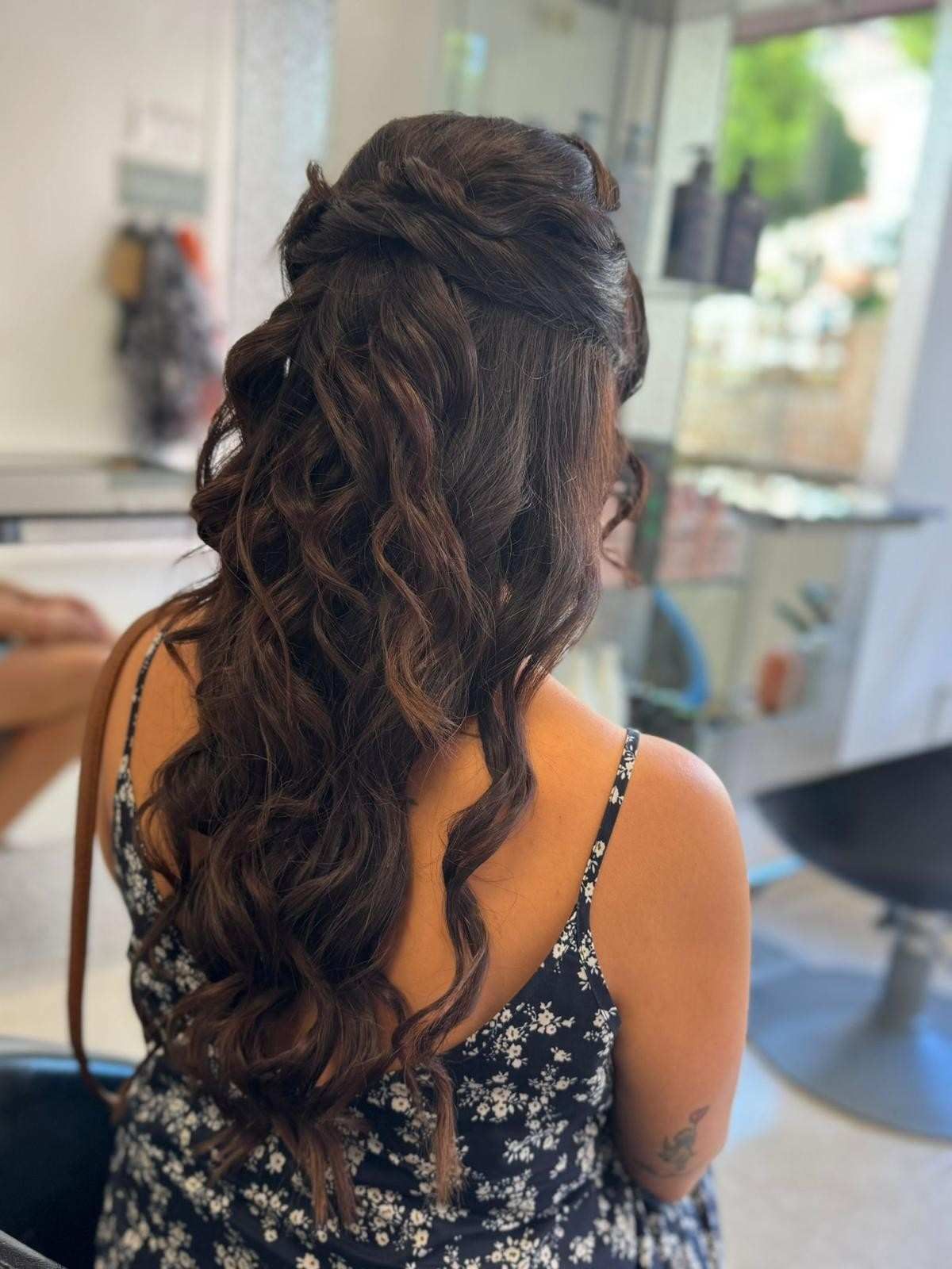 Mujer con cabello castaño largo y ondulado, recogido hasta la mitad. Lleva un vestido con estampado floral en un salón de belleza.