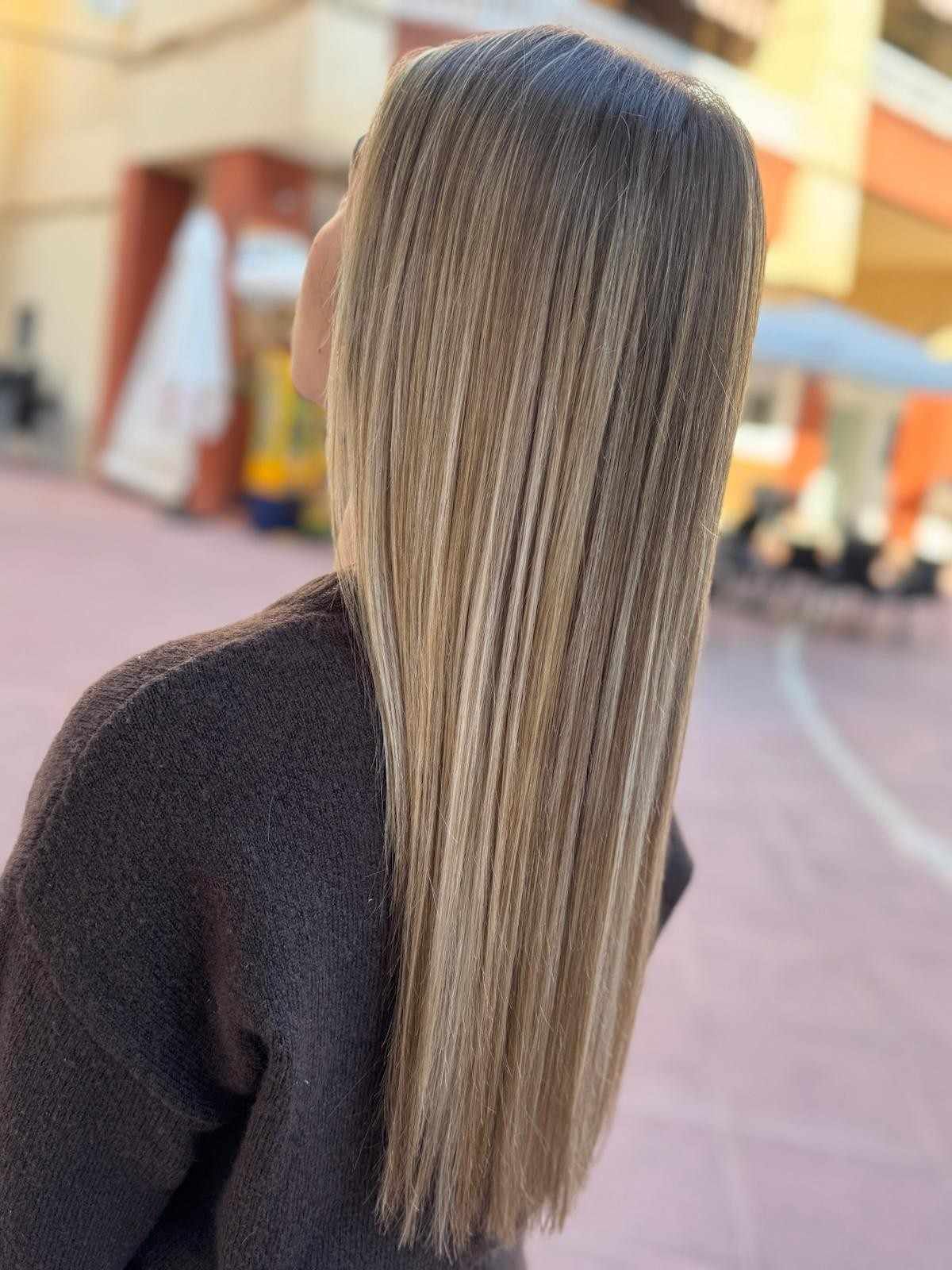 Mujer con cabello rubio largo y rizado, de espaldas a la cámara, vistiendo un suéter marrón, al aire libre.