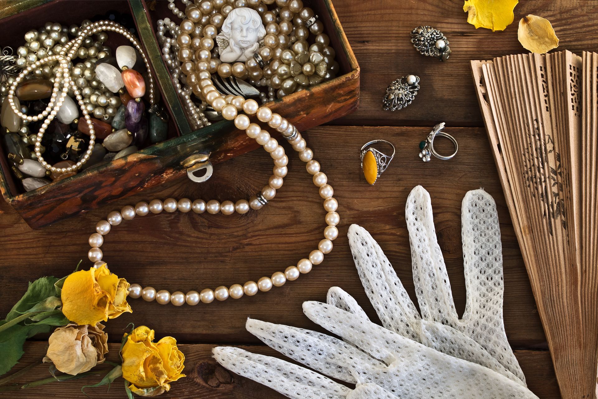 Boîte à bijoux en bois contenant des perles et autres bijoux, un éventail, des gants et des fleurs jaunes posés sur une surface en bois.