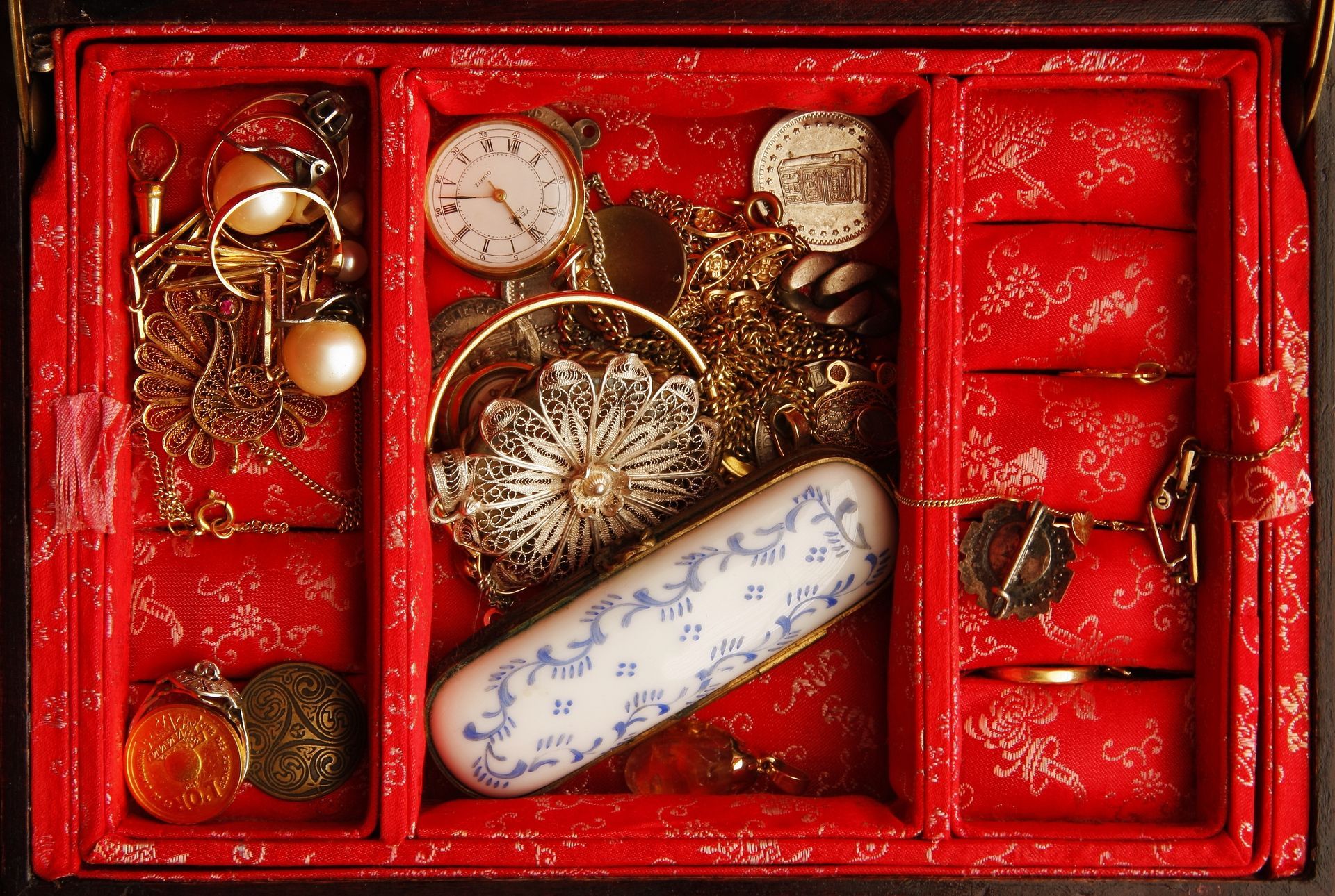 Boîte à bijoux rouge ouverte, remplie de bijoux assortis en or et en argent, dont une montre et des pièces de monnaie.