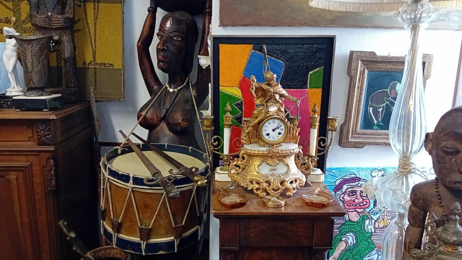 Vitrine d'un magasin d'antiquités présentant un tambour, des statues, une horloge et des œuvres d'art.