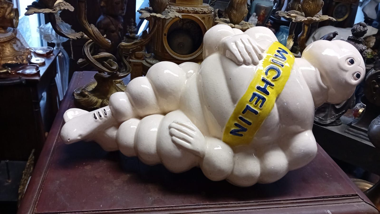 Figurine de Bibendum allongée, blanche avec une bannière jaune, placée au milieu d'autres antiquités.