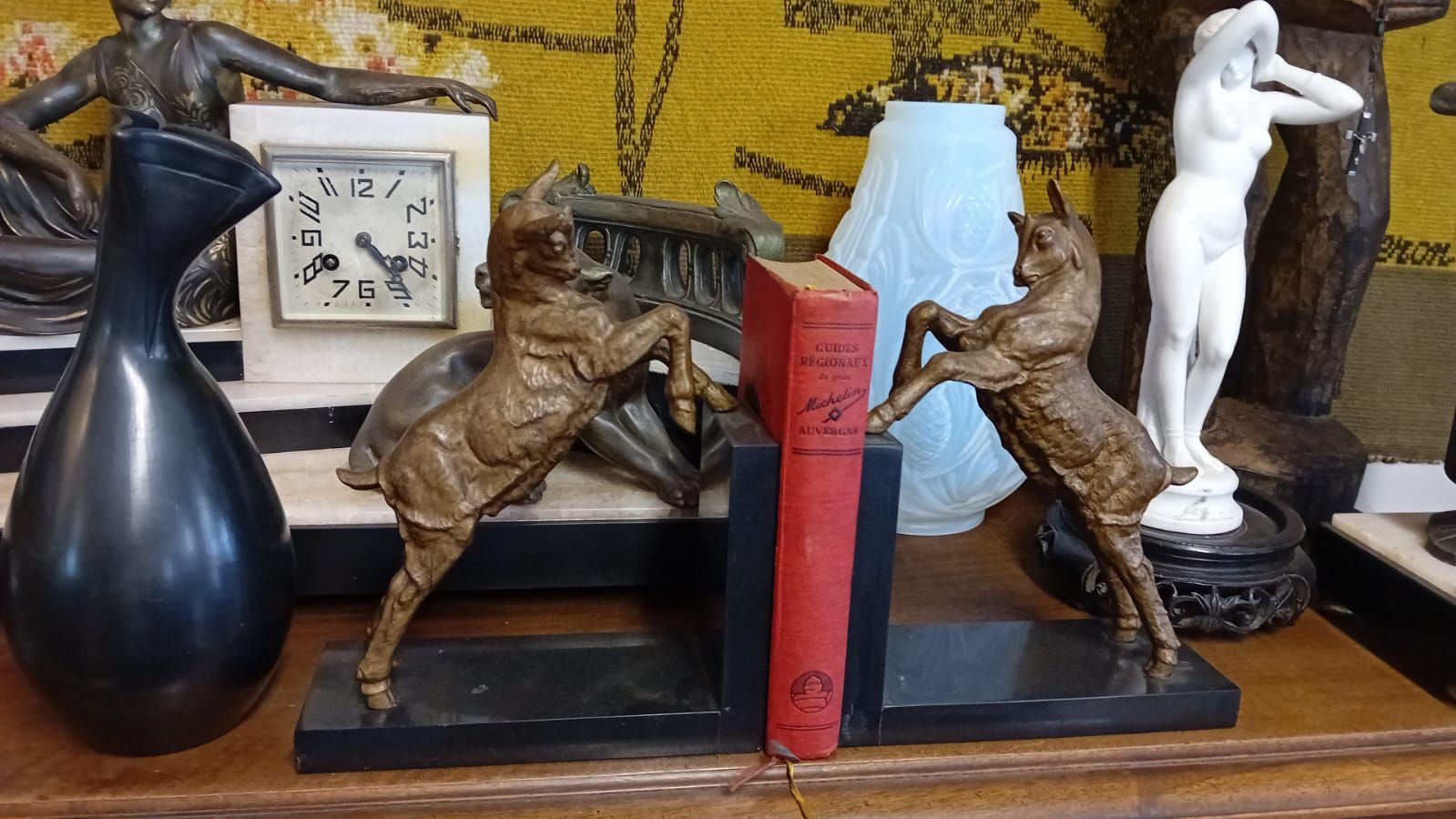 Des livres maintenus par des serre-livres en forme de chèvre en bronze, posés sur une étagère avec des objets décoratifs.
