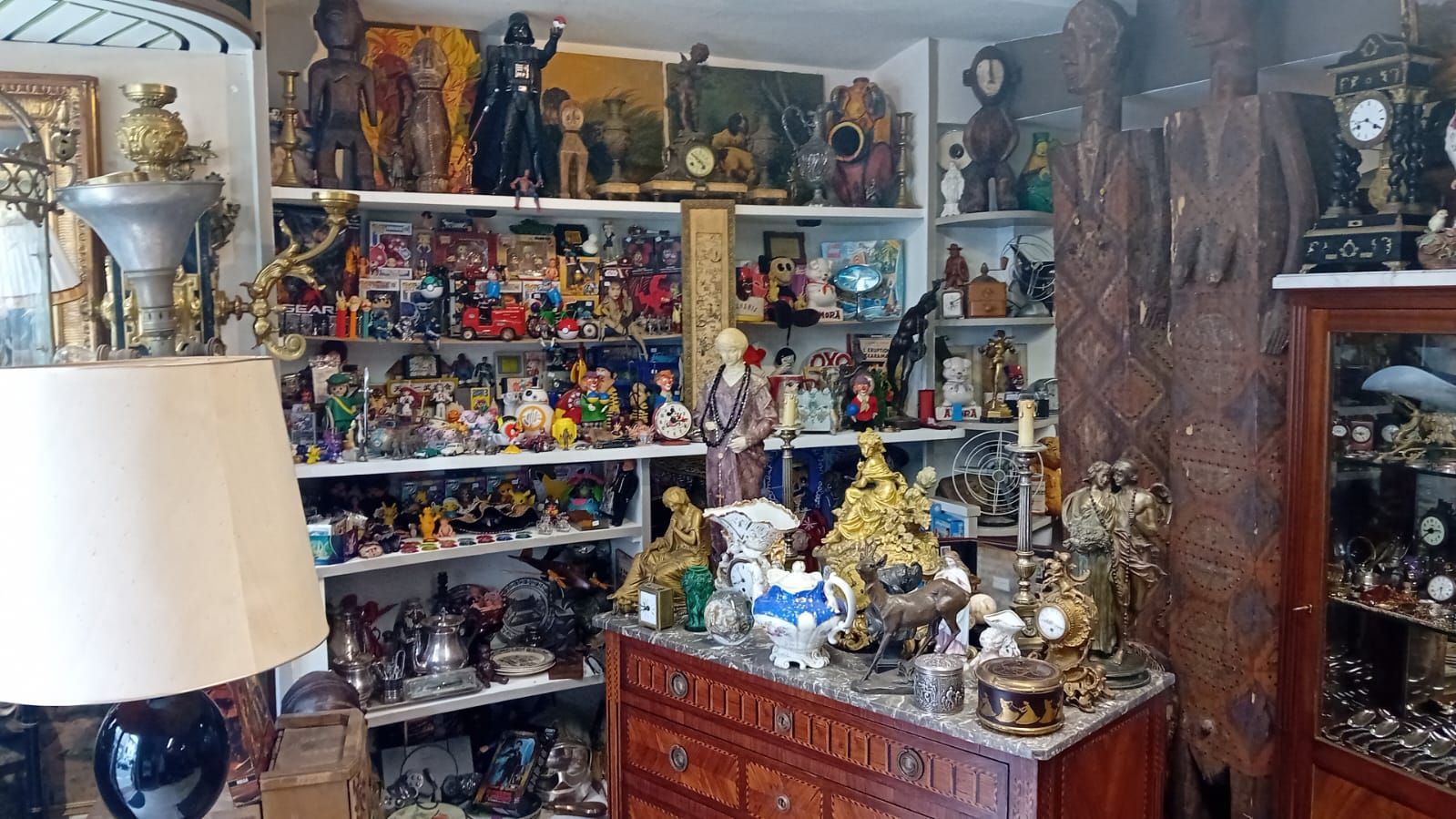 Une pièce remplie d'objets anciens divers, notamment des sculptures, des horloges et des figurines, exposés sur des étagères et des meubles.