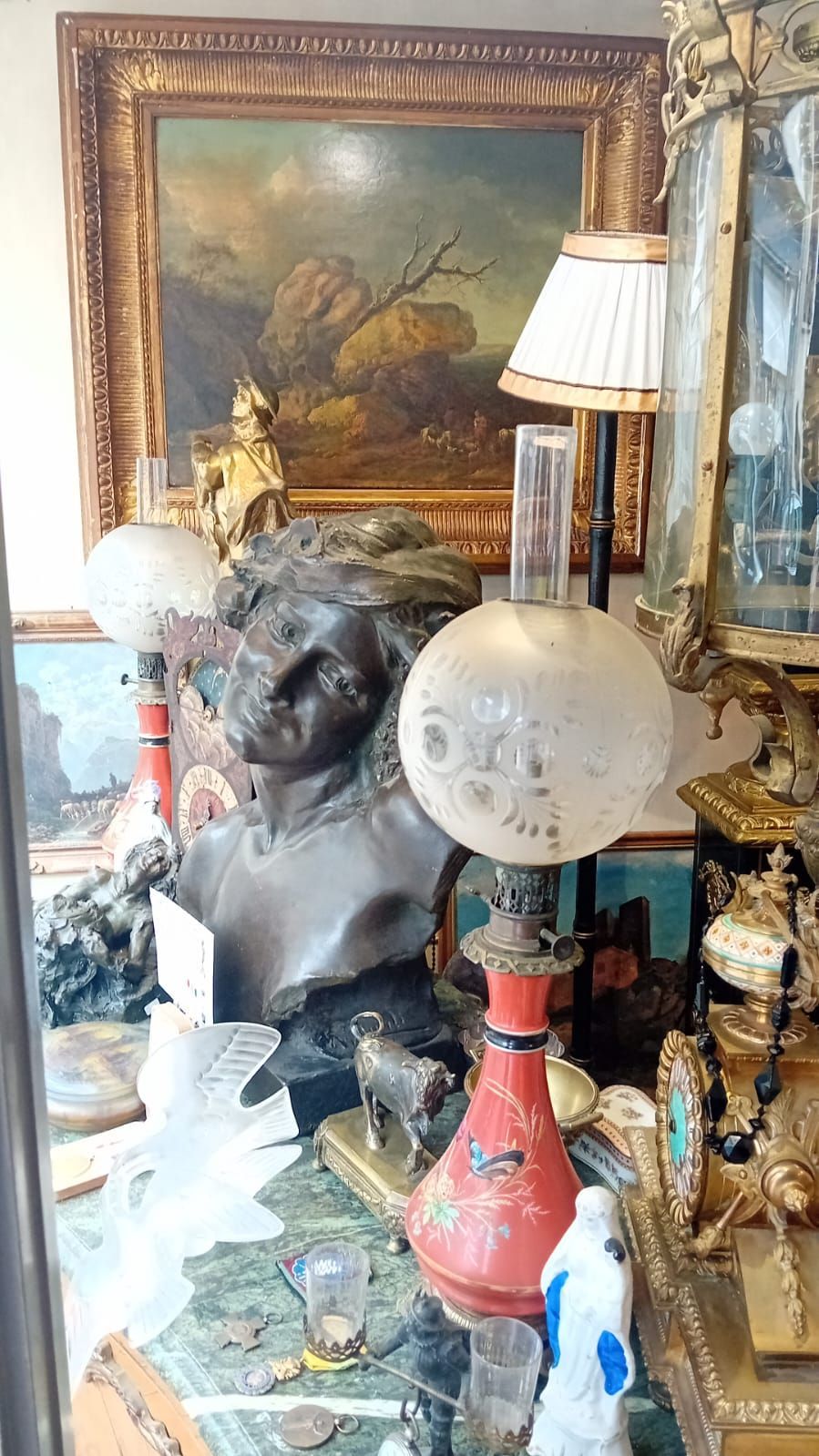 Vitrine d'antiquaire : buste, lampes, tableau et objets ornementaux disposés sur une table. Teintes or et rouge.
