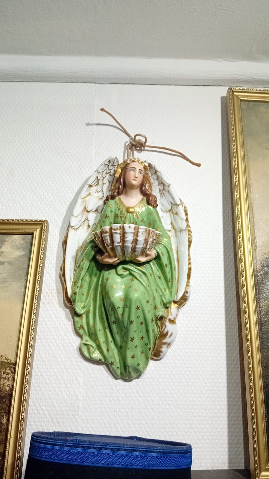 Figurine d'ange en robe verte, tenant un panier, fixée au mur entre deux tableaux encadrés.