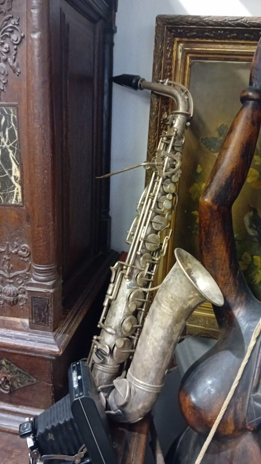 Un saxophone argenté appuyé contre un meuble en bois sombre, près d'un tableau encadré et de meubles en bois.