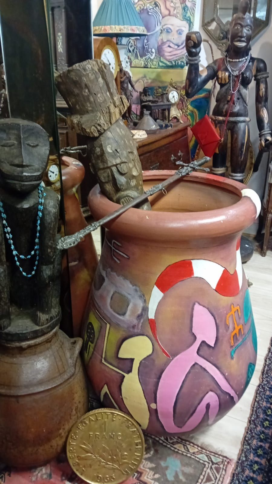 Un grand pot coloré orné d'illustrations et d'une pièce de monnaie sur le devant, entouré de figurines en bois sculpté et d'antiquités.