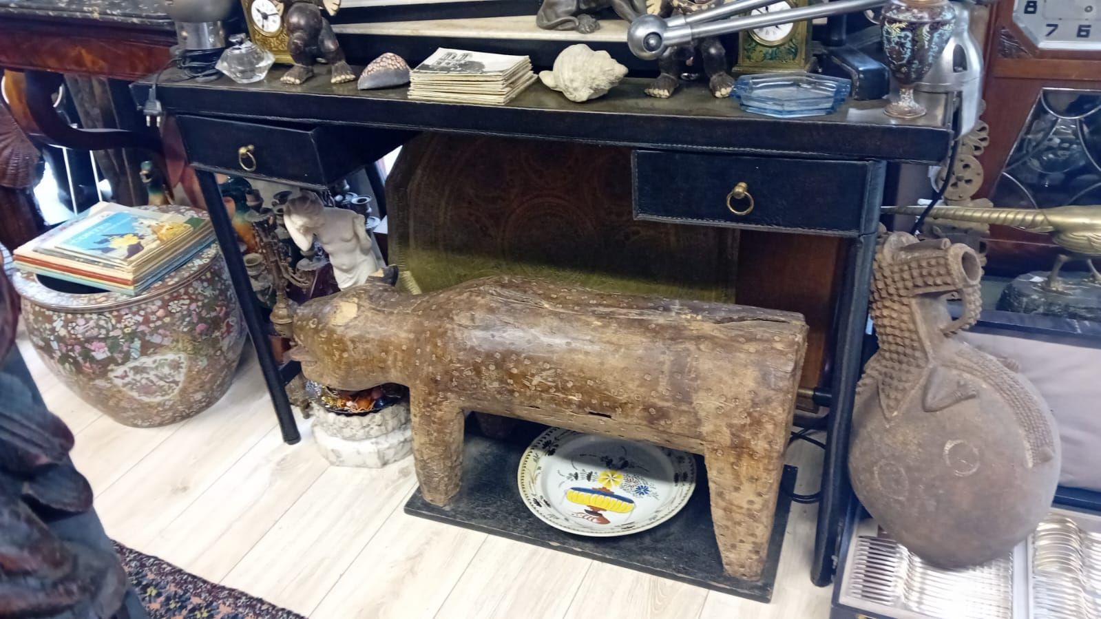 Une boutique d'antiquités encombrée : un bureau noir à tiroirs, un banc en pierre, divers objets décoratifs et un récipient à motifs partiellement visible.