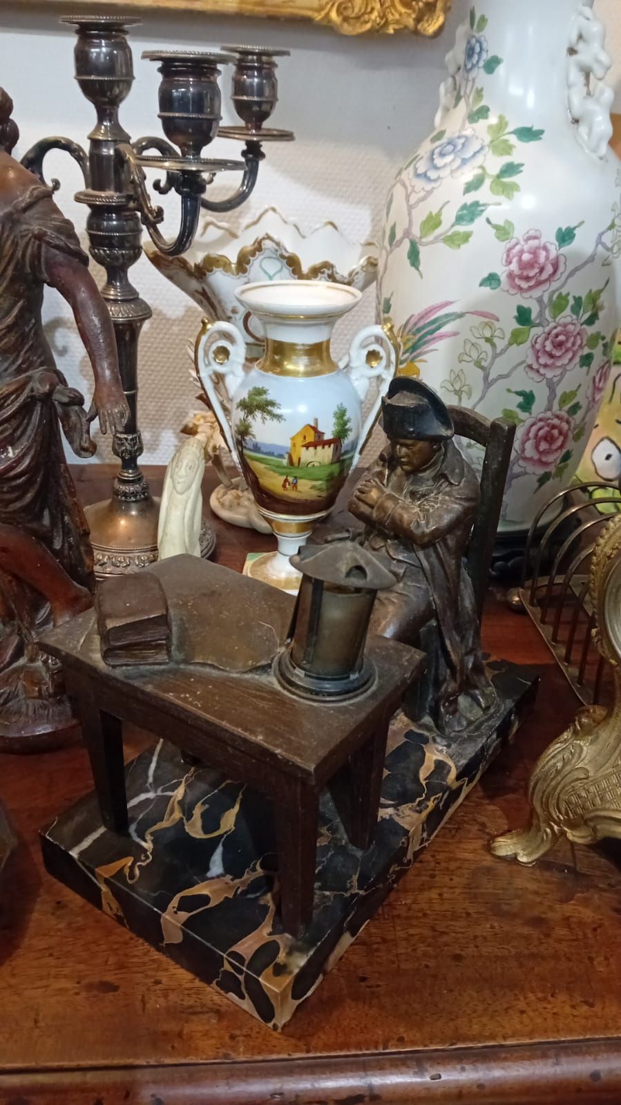 Sculpture en bronze représentant un personnage assis à un bureau avec une lampe, entouré d'objets décoratifs anciens.