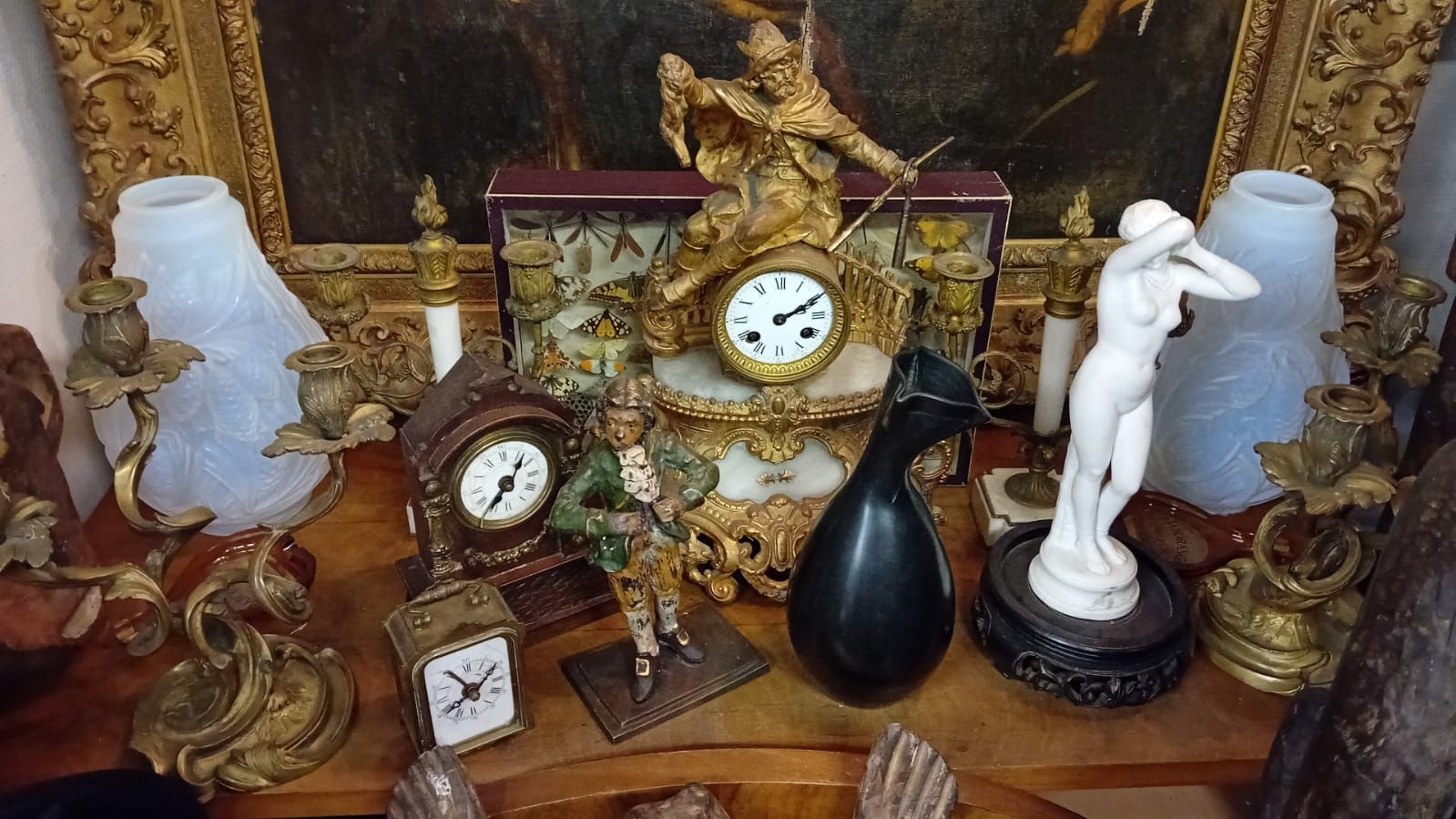 Une collection d'horloges anciennes, de candélabres, de sculptures et d'œuvres d'art disposées sur une surface en bois, avec un tableau en arrière-plan.