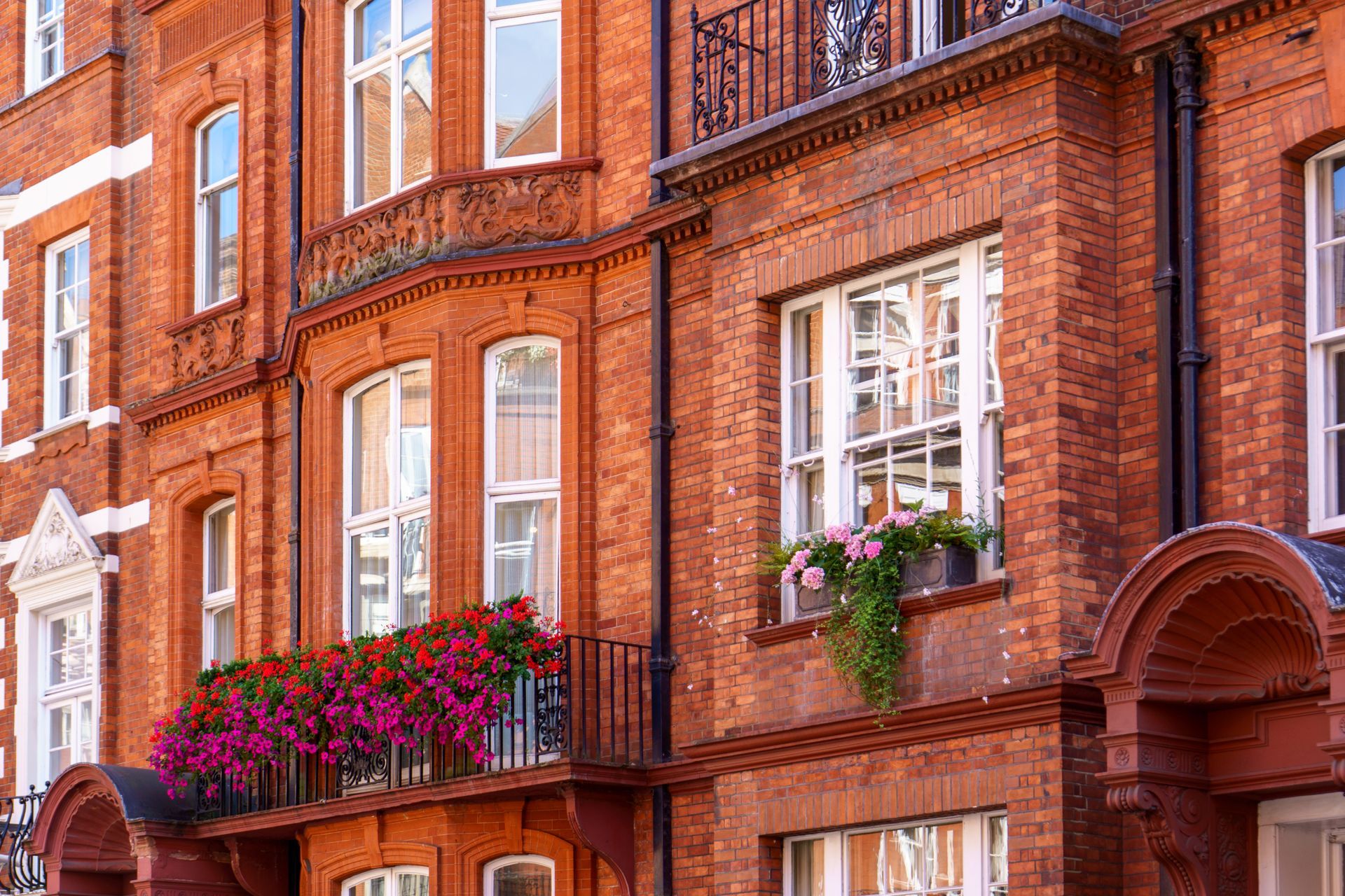 Façades de maisons de ville en briques rouges avec fenêtres à guillotine, balcons en fer noir et jardinières débordantes de fleurs.