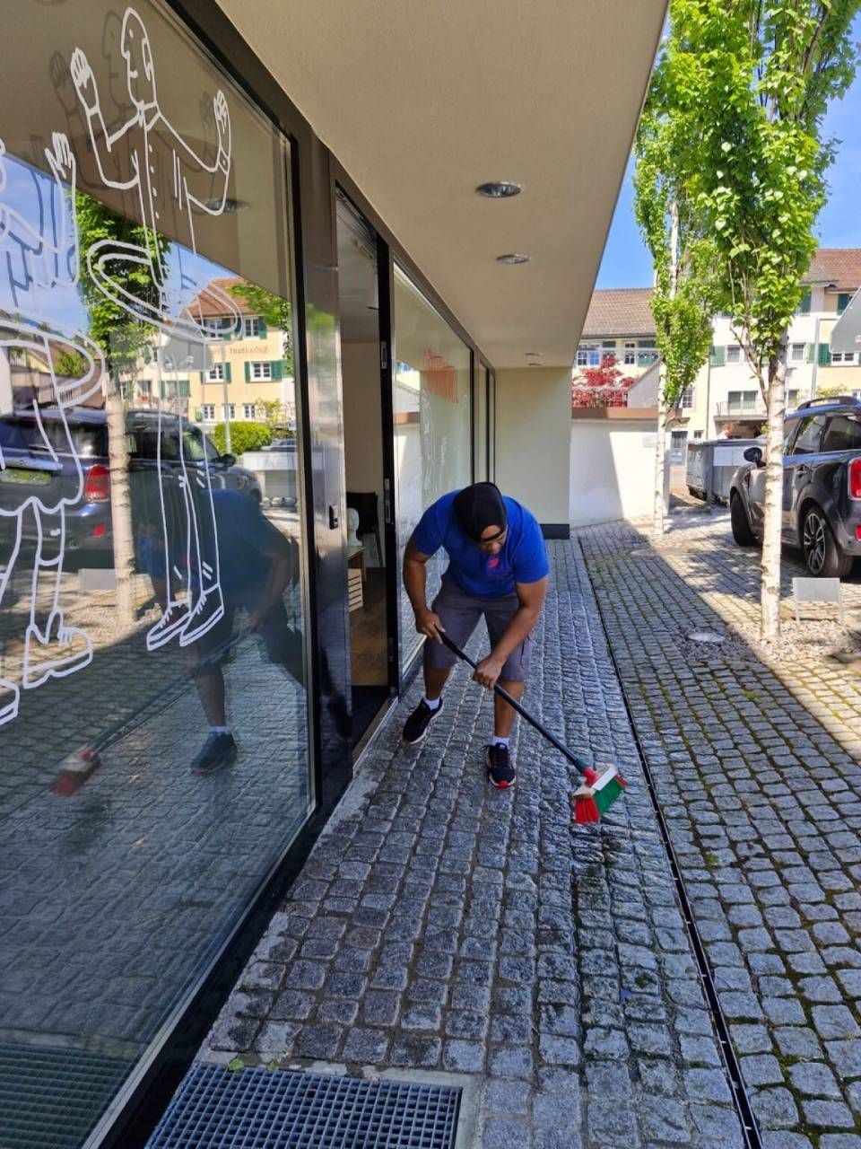 Ein Mann fegt unter sonnigem Himmel einen Kopfsteinpflasterweg neben einem Schaufenster mit Fensteraufklebern.