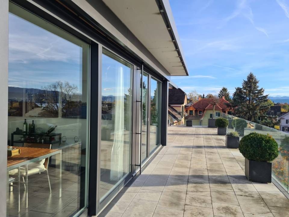 Balkon mit Glasschiebetüren, Steinboden, Topfpflanzen und Blick auf Häuser und Bäume an einem sonnigen Tag.