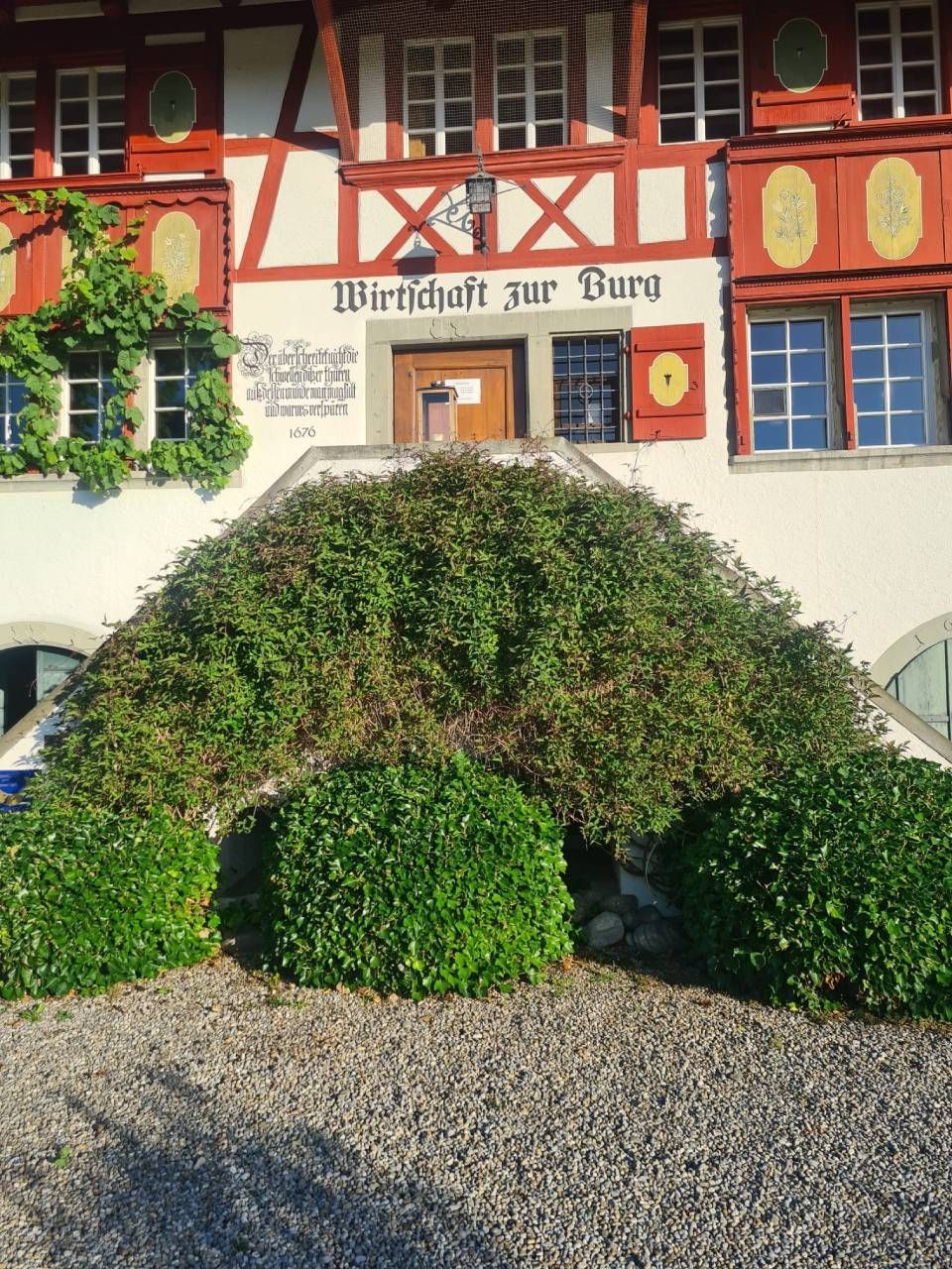 Gebäude mit rot-weisser Fassade, Schild „Wirtschaft zur Burg“, üppiges Grün über der Treppe und rund um den Gebäudeeingang.