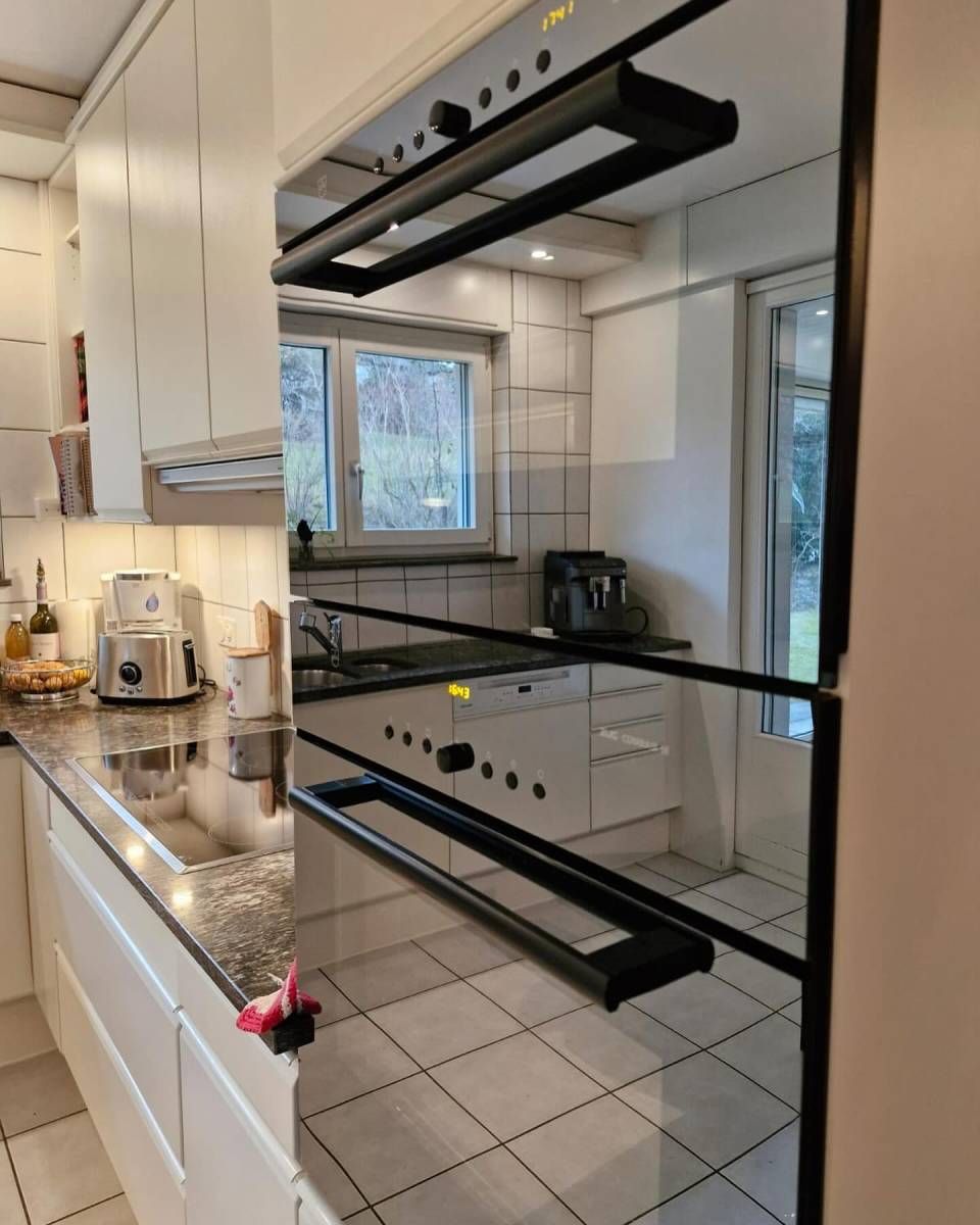 Küche mit weissen Schränken, dunkler Arbeitsplatte und eingebautem schwarzem Backofen, der den Raum reflektiert.