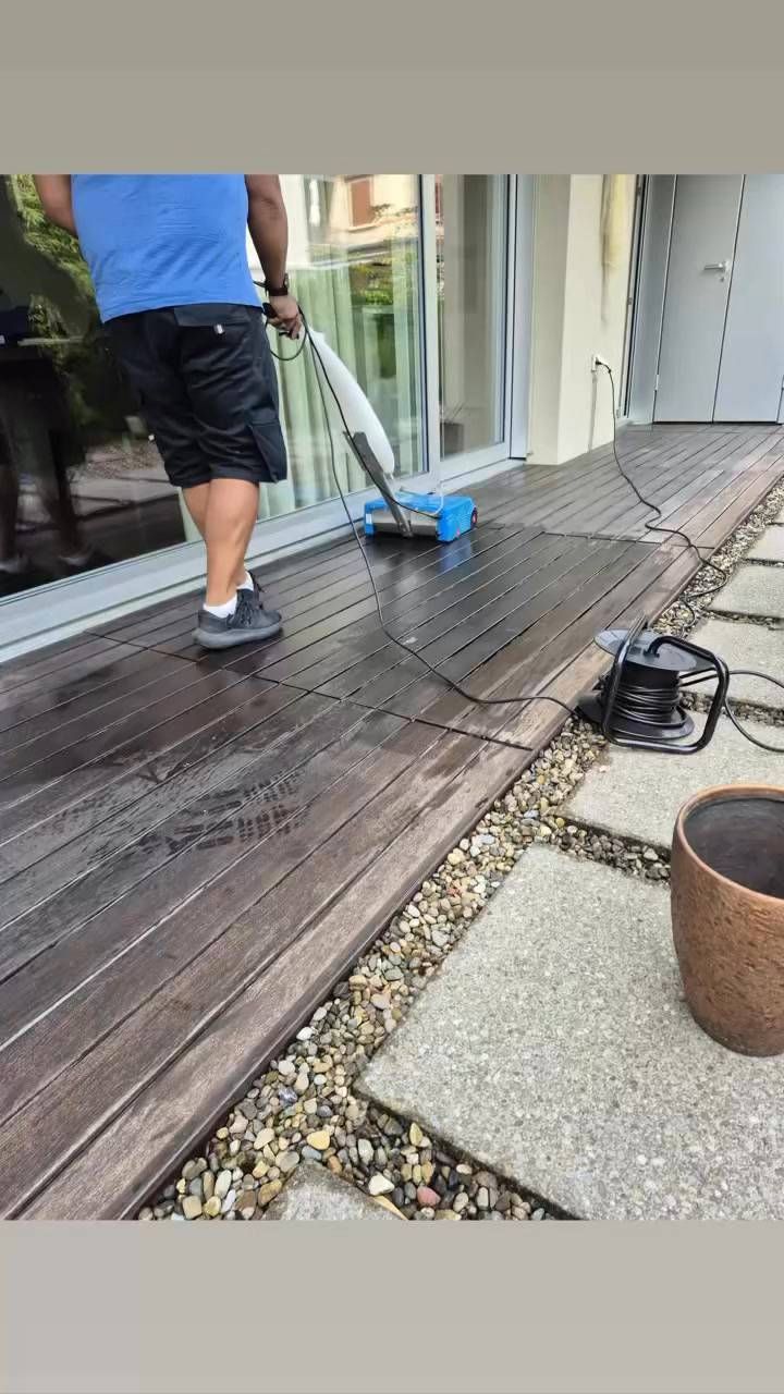 Eine Person reinigt mit einer Reinigungsmaschine eine nasse Holzterrasse in der Nähe eines Gebäudes, von Steinen und einem Topf.