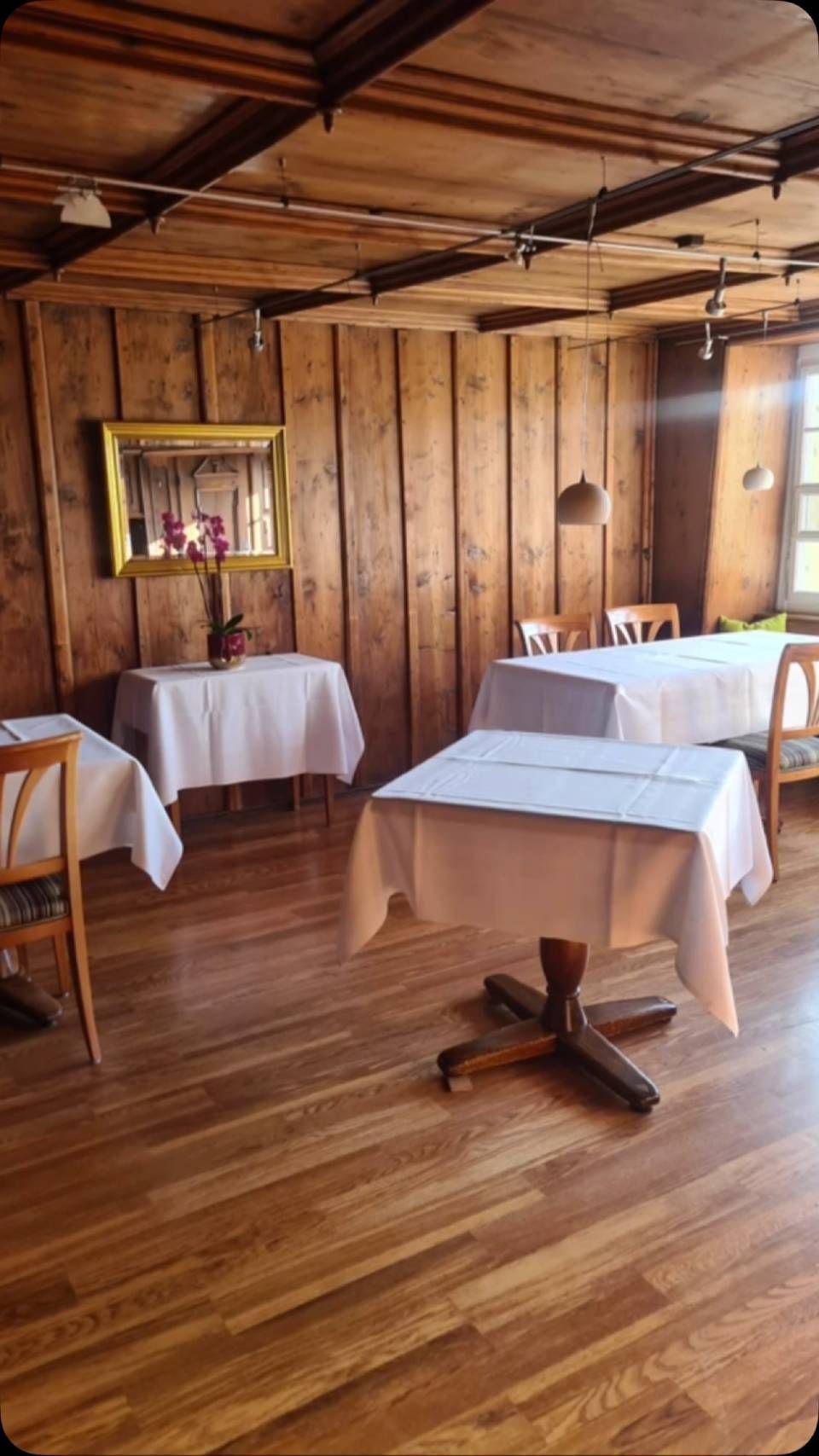 Innenansicht eines Restaurants mit holzgetäfelten Wänden, Tischen mit weissen Tischdecken und Holzböden.