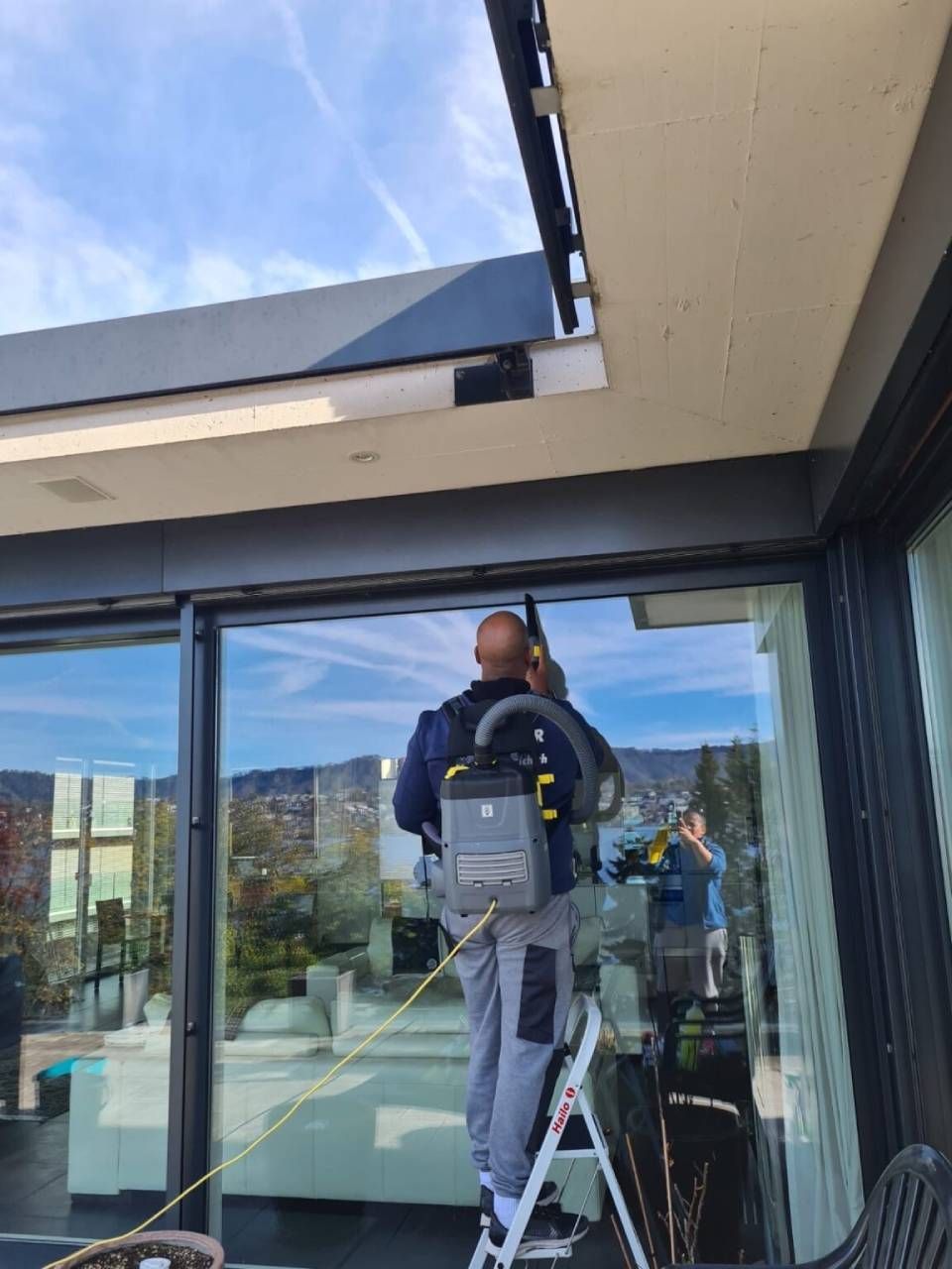 Person putzt Fenster auf einer Leiter mit einem Rucksackstaubsauger; sonniger Tag mit Bergblick.