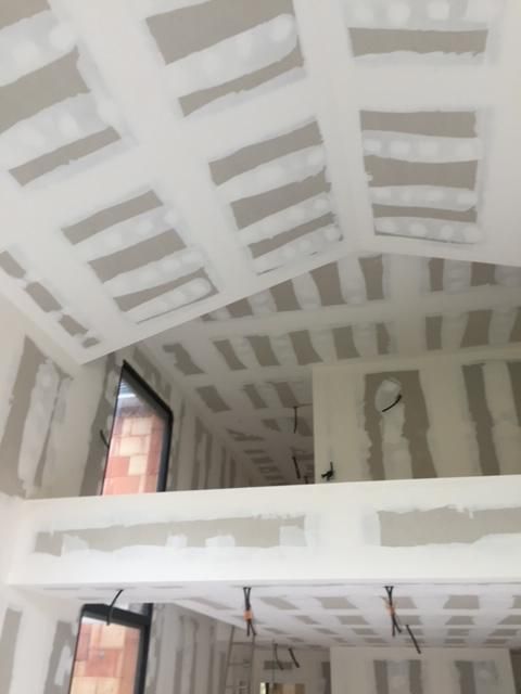 Intérieur d'un bâtiment en construction, avec des plaques de plâtre installées sur les murs et le plafond ; câblage électrique visible.