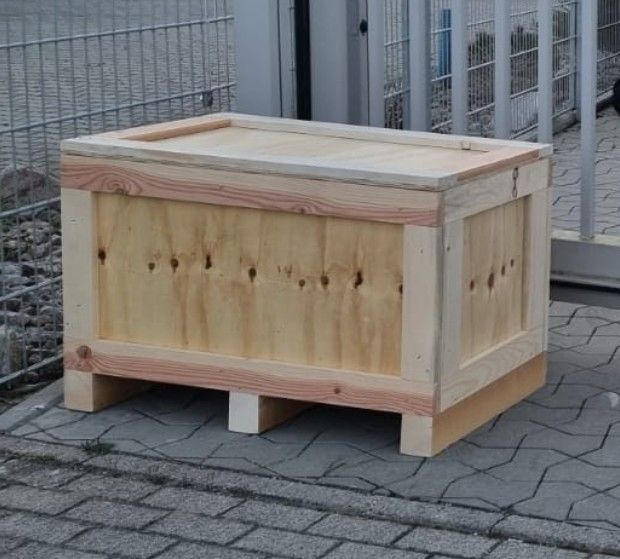 Auf einem gepflasterten Gehweg neben einem Zaun steht eine Holzkiste.