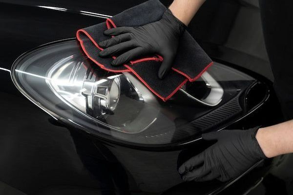 Una persona que lleva guantes negros limpia un faro de un coche con un paño de microfibra negro y rojo.