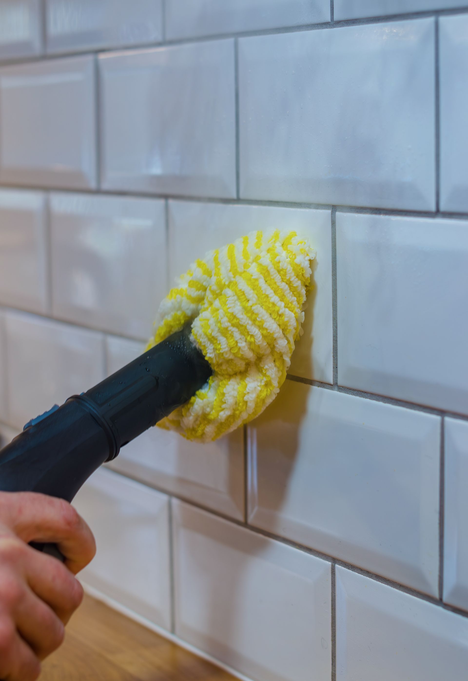 Personne utilisant un nettoyeur vapeur avec un tampon jaune pour nettoyer un mur carrelé de carreaux de métro blancs.