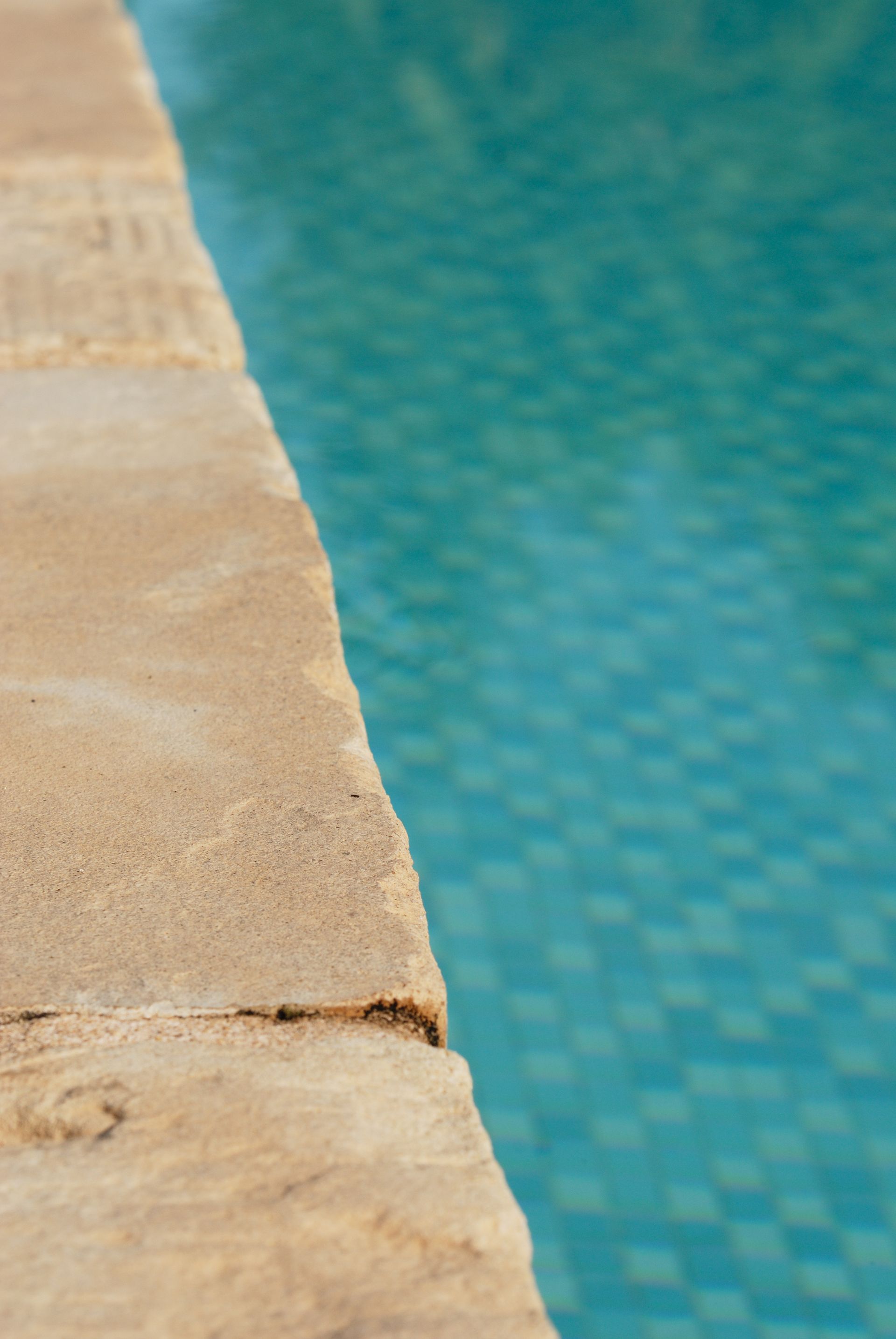 Bord de piscine en pierre beige, bordée d'une eau turquoise et carrelée de petits carreaux carrés.