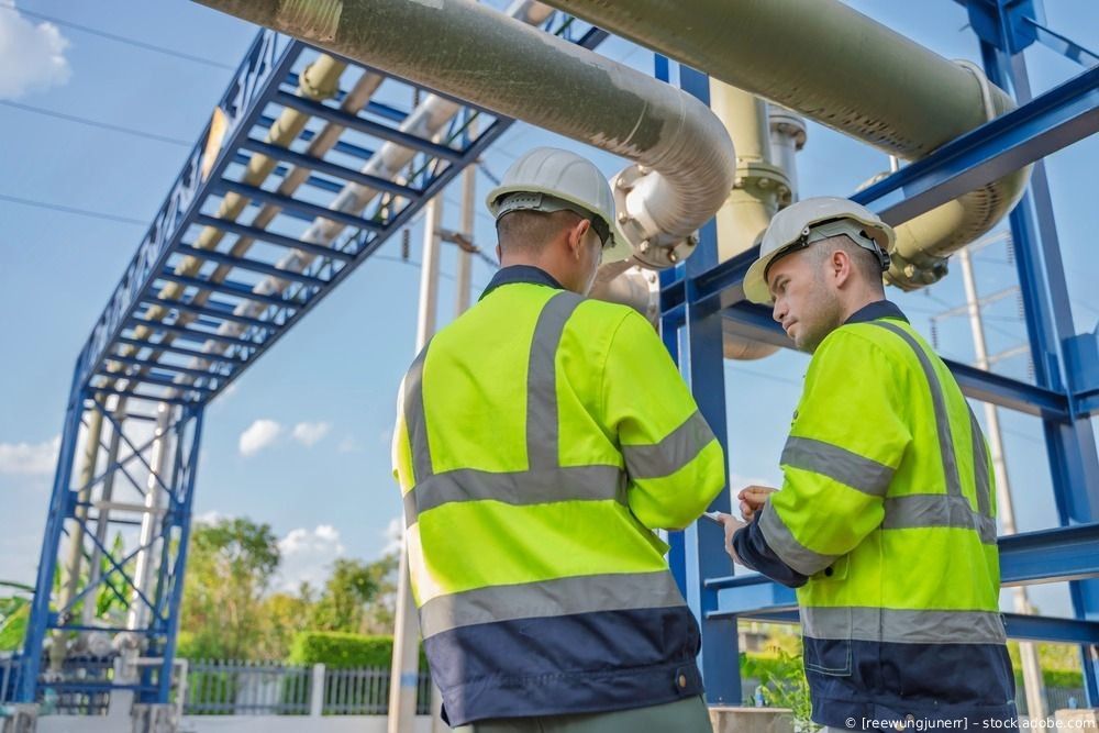Zwei Arbeiter in neonfarbenen Sicherheitswesten inspizieren Industrierohre im Freien unter blauem Himmel
