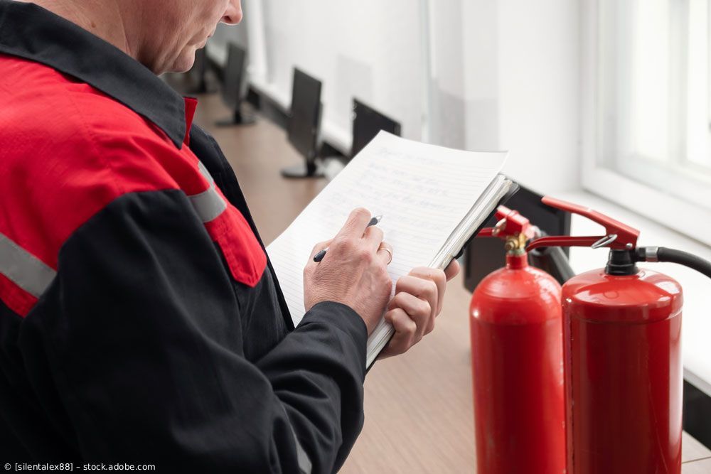 Ein Techniker schreibt auf einem Klemmbrett neben zwei roten Feuerlöschern in einem hellen Büro.