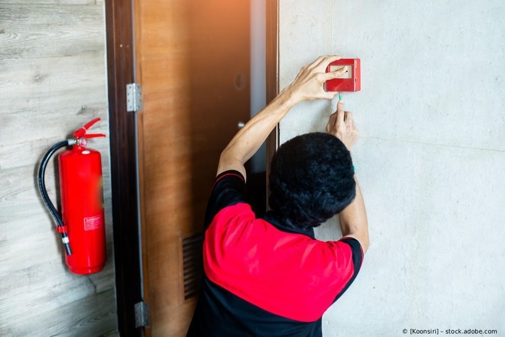 Eine Person montiert einen roten Feuermelder an einer Wand neben einem Feuerlöscher.