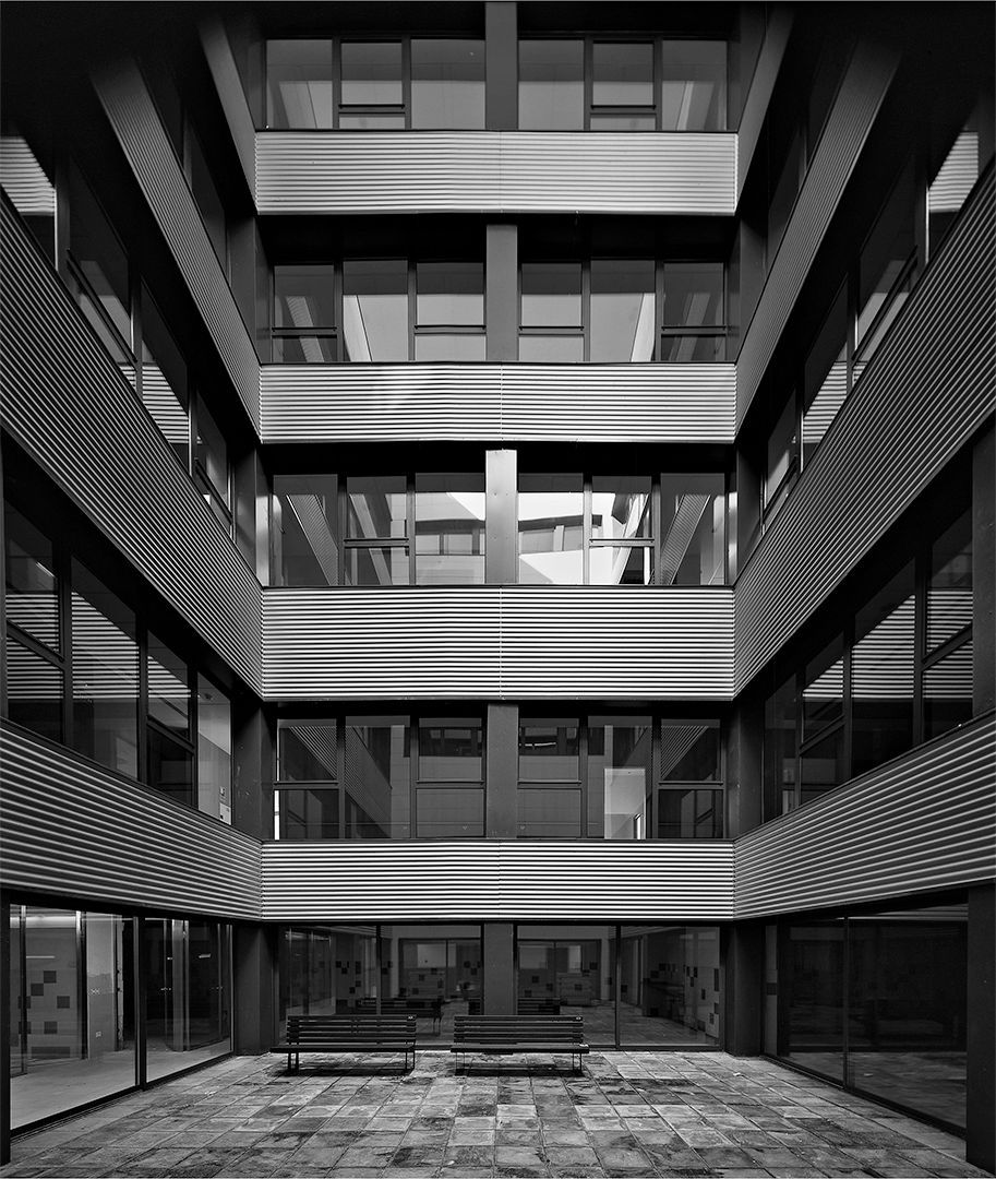 Patio de edificio moderno, de varios pisos, con ventanas reflectantes y bancos en el suelo.