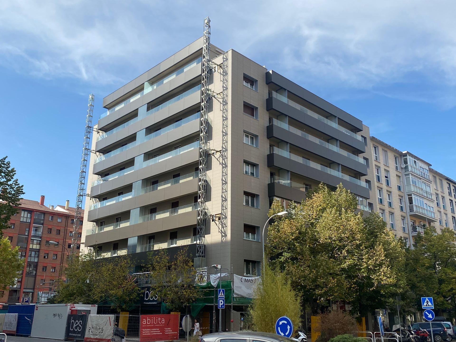 Edificio moderno con fachada marrón y plata, rodeado de árboles y barreras de construcción.