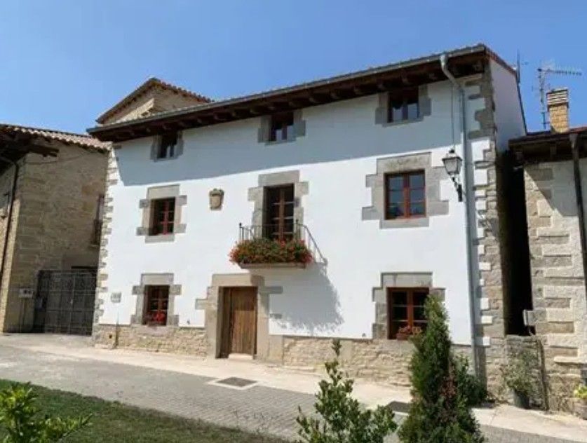 Edificio blanco de dos pisos con detalles en piedra, puerta y ventanas de madera marrón, balcón con flores.